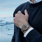 Grey watch with an orange dial outline worn on a raised wrist beside a navy sweater and snowy landscape.