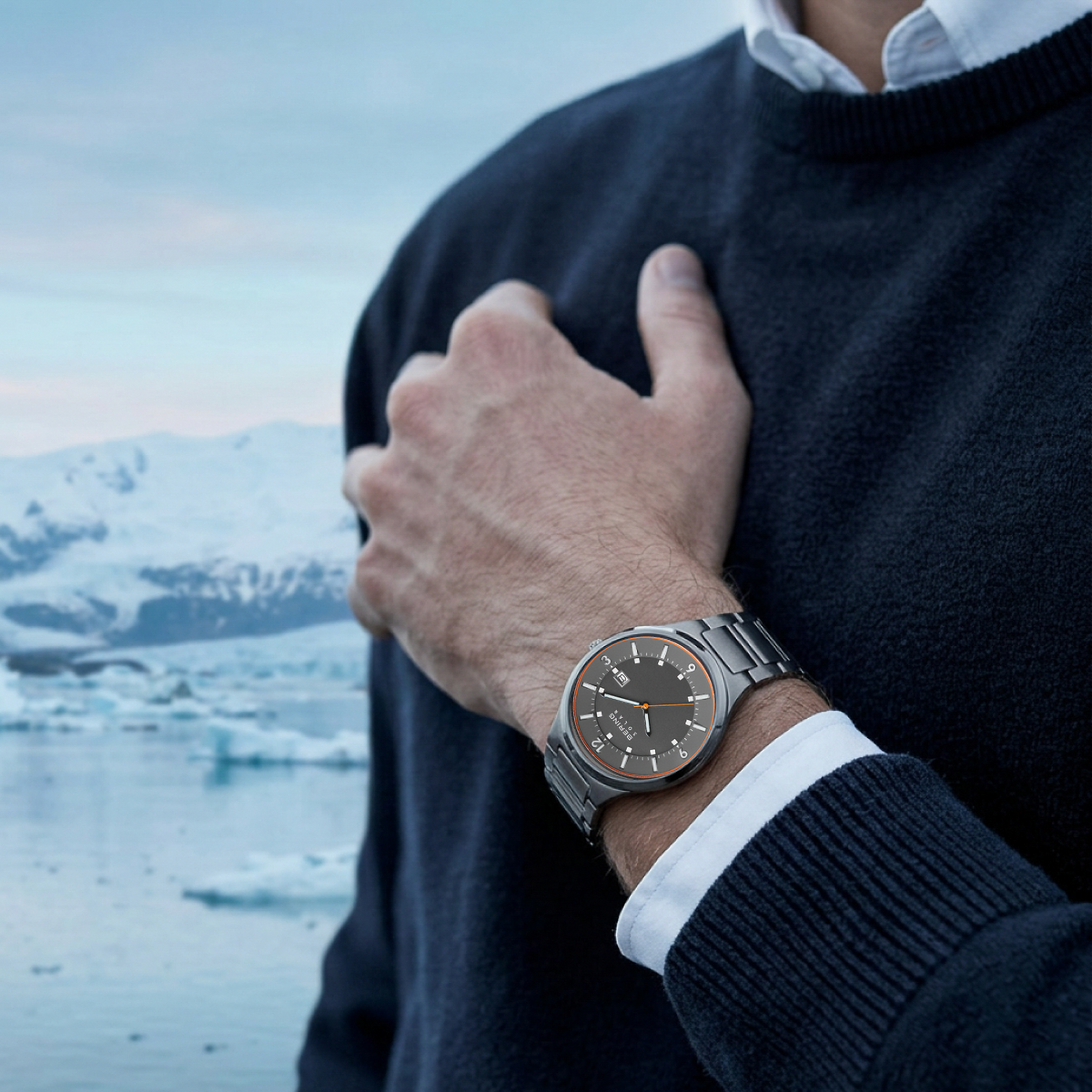Grey watch with an orange dial outline worn on a raised wrist beside a navy sweater and snowy landscape.