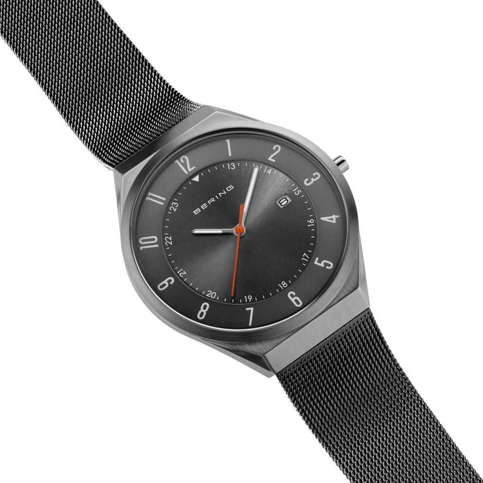 Diagonal overhead view of the grey dial with an orange hand, grey case and grey mesh strap.