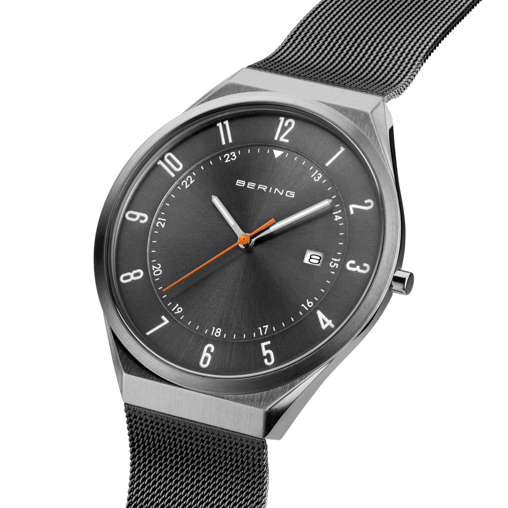Angled close-up of the grey dial with an orange hand and grey case beside the grey mesh strap.